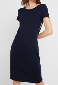 Marinblå klänning med kort ärm i mjukt material, figurnära design, med sidodekor som har textmönster, knä-lång fåll och rund halsringning.