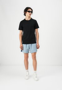 T-shirt noir en coton, shorts bleu clair avec bandes latérales, baskets blanches et lunettes de soleil. Le modèle se tient contre un fond blanc.