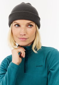 Jeune femme aux cheveux blonds portant un bonnet en tricot gris et un sweat à capuche en polaire teal avec le logo "protest", regardant légèrement sur le côté.