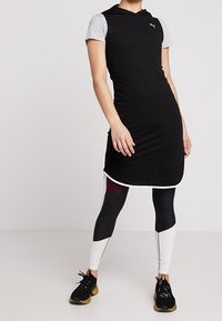 Svart sportklänning med korta grå ärmar, huva och vita detaljer; kombinerad med flerfärgade leggings och svarta träningsskor.