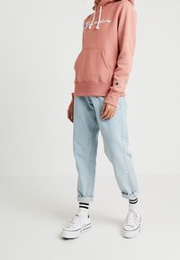 Vaaleanpunainen huppari valkoisilla kirjaimilla ja etutaskulla, yhdistetty vaaleansinisiin farkkuihin, jotka on käännetty lahkeista ylös. Valkoiset lenkkarit mustavalkoisilla raitasukilla.