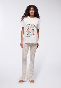 T-shirt bianco in cotone con grafica pesca, abbinato a pantaloni leggeri in maglia crema con motivo a diamante, indossati con infradito beige.