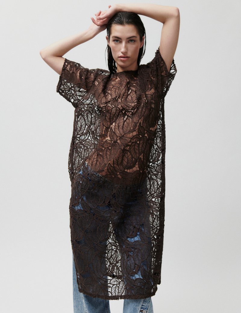 Γυναίκα που ποζάρει με τα χέρια σηκωμένα, φορώντας μακρύ καφέ δαντελωτό τοπ πάνω από μπλε τζιν, με απλό ανοιχτό φόντο.