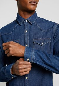 Dunkelblauses Denimhemd mit spitzem Kragen, langen Ärmeln, zwei Brusttaschen und kontrastierenden Nähten. Verfügt über eine Knopf-Front und metallische Knöpfe.