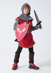 Child dressed as a knight, wearing a red tunic with gold trim, gray chainmail hood, brown boots, and holding a pink shield and sword.