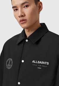 Giacca nera con bottoni a pressione, caratterizzata da un logo con simbolo di pace e testo "ALLSAINTS" in bianco. Tessuto liscio con design del colletto.