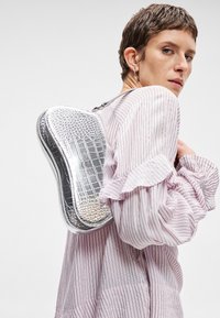 Ασημένιο σακίδιο με ανάγλυφο σχέδιο κροκοδείλου και κομψό, στρογγυλεμένο σχήμα. Διαθέτει γυαλιστερή επιφάνεια και μια ενιαία θήκη που φοριέται πάνω από ένα ροζ πουκάμισο με ρίγες.