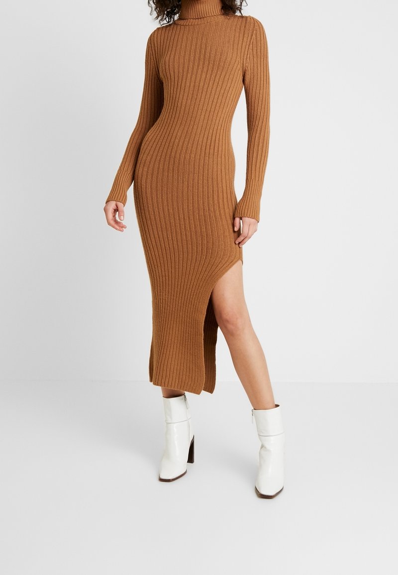 Femme en robe midi marron côtelée ajustée à col roulé avec une fente latérale, portant des bottines blanches à talons, debout devant un fond uni.