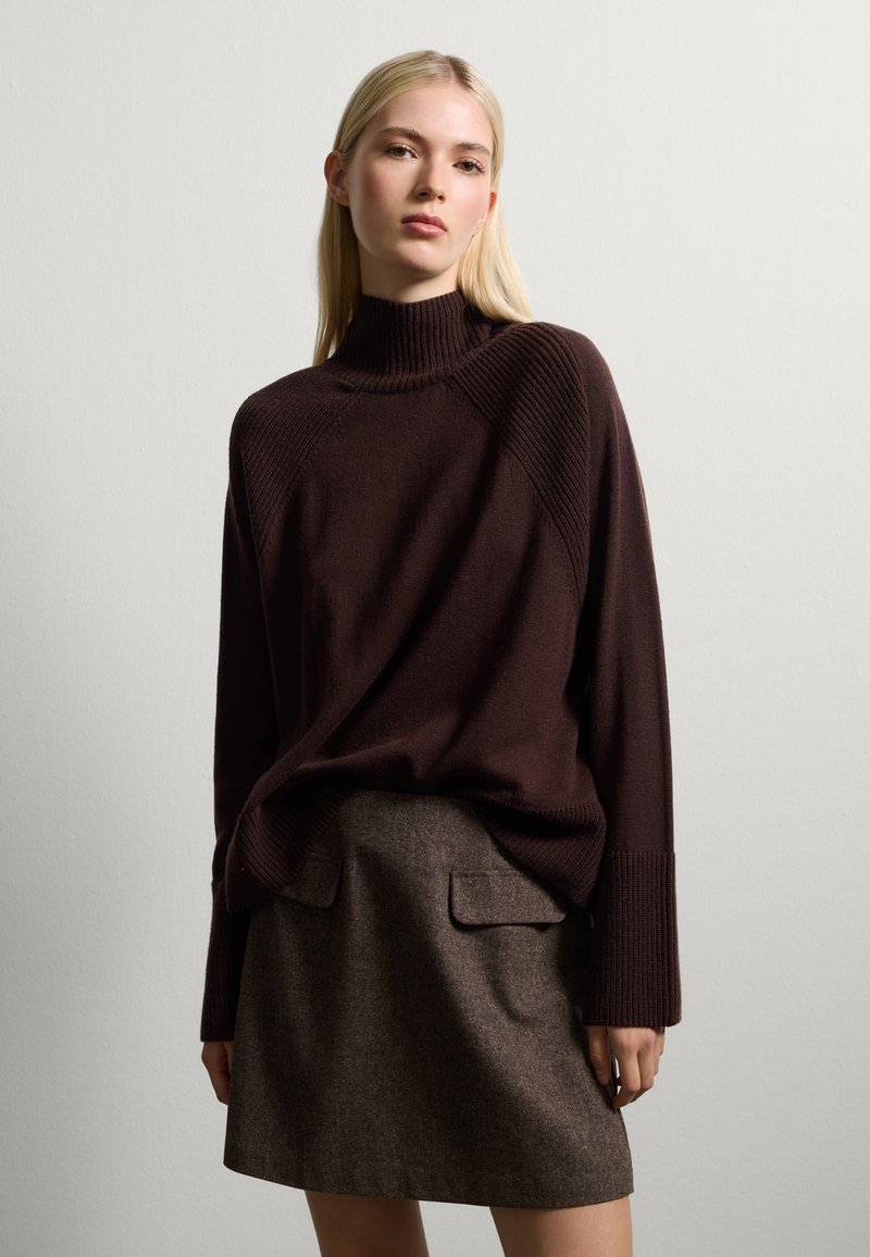 Un pull marron foncé avec un col cylindrique haut, des manches texturées et une coupe ample, associé à une jupe marron avec des poches plaquées.