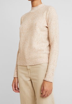 Person iført en beige strikket sweater med ribbet manchetter og kant, kombineret med lyse beige bukser, mod en ensfarvet grå baggrund.