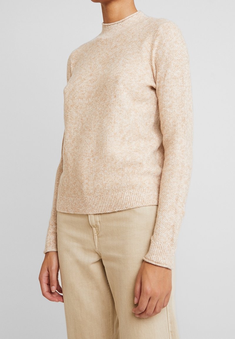 Personne portant un pull tricoté beige avec poignets et ourlet côtelés, associé à un pantalon beige clair, sur un fond gris uni.