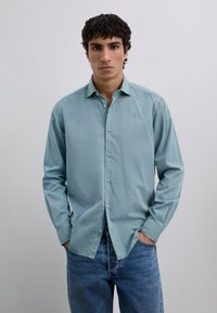 Lichtblauw overhemd met knopen en kraag, lange mouwen en subtiele borduursels op de borst, gecombineerd met donkerblauwe spijkerbroek.