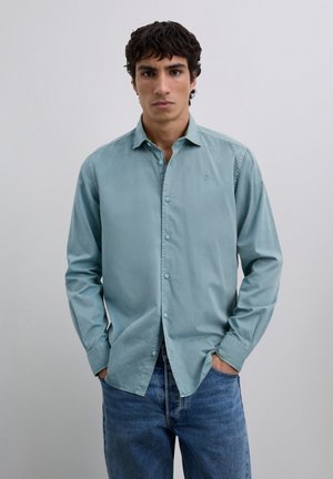 Camicia azzurro chiaro con bottoni, colletto, maniche lunghe e ricamo delicato sul petto, abbinata a jeans blu scuro.
