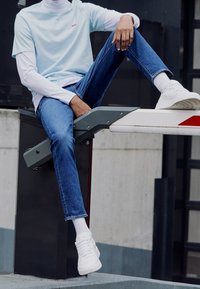 Camiseta azul claro con logo rojo, superpuesta a una camisa de manga larga blanca. Jeans azules y zapatillas blancas; sentado sobre una estructura gris.