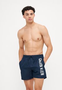 Marinefarbene Schwimmshorts mit elastischem Bund und Kordelzug, ausgestattet mit einem weißen "CALVIN KLEIN"-Logo, das vertikal auf der linken Seite aufgedruckt ist.