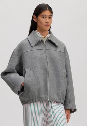 Vrouw draagt een grijze wollen jas met rits, wijde mouwen en elastische zoom over een gestreepte broek, staand met één hand in de zak.