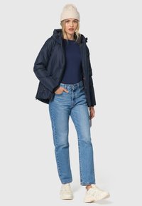 Marineblauer gepolsterter Mantel mit Kapuze über einem marineblauen Oberteil, kombiniert mit hellblauen Jeans und beigen Sneakers. Das Model trägt eine cremefarbene Strickmütze.