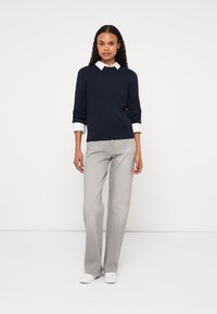 Jūras spēku krāsas adīta džemperis ar teksturētu rakstu, balta kreklu apakšā, apvienots ar gaišiem pelēkiem taisnām kājām džinsiem un baltiem sporta apaviem.