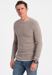 Maglione lungo beige chiaro a maniche lunghe con un motivo testurizzato, scollatura rotonda e bordi bianchi. Indossato con jeans blu e un orologio con dettagli marroni.