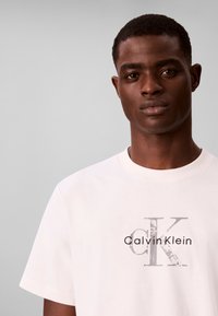 Νέος άνδρας με κοντά μαλλιά φορώντας λευκό t-shirt Calvin Klein σε απλό ανοιχτό γκρι φόντο.