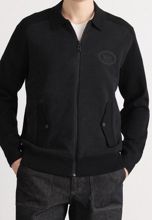 Schwarze Reißverschlussjacke mit Kragen und vorderen Klappentaschen, getragen über einem weißen Hemd, kombiniert mit dunklen Denim-Jeans.