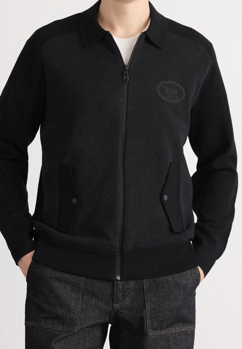 Veste noire zippée avec col et poches avant à rabat, portée sur une chemise blanche, associée à un jean en denim foncé.