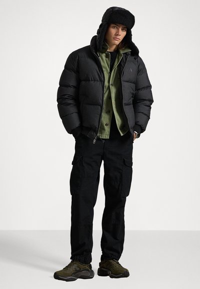 Čierna nadýchaná bunda s plyšovou kapucňou, zelená košeľa s gombíkmi pod ňou, čierne cargo nohavice a tmavé športové topánky. Neformálny outdoorový štýl.