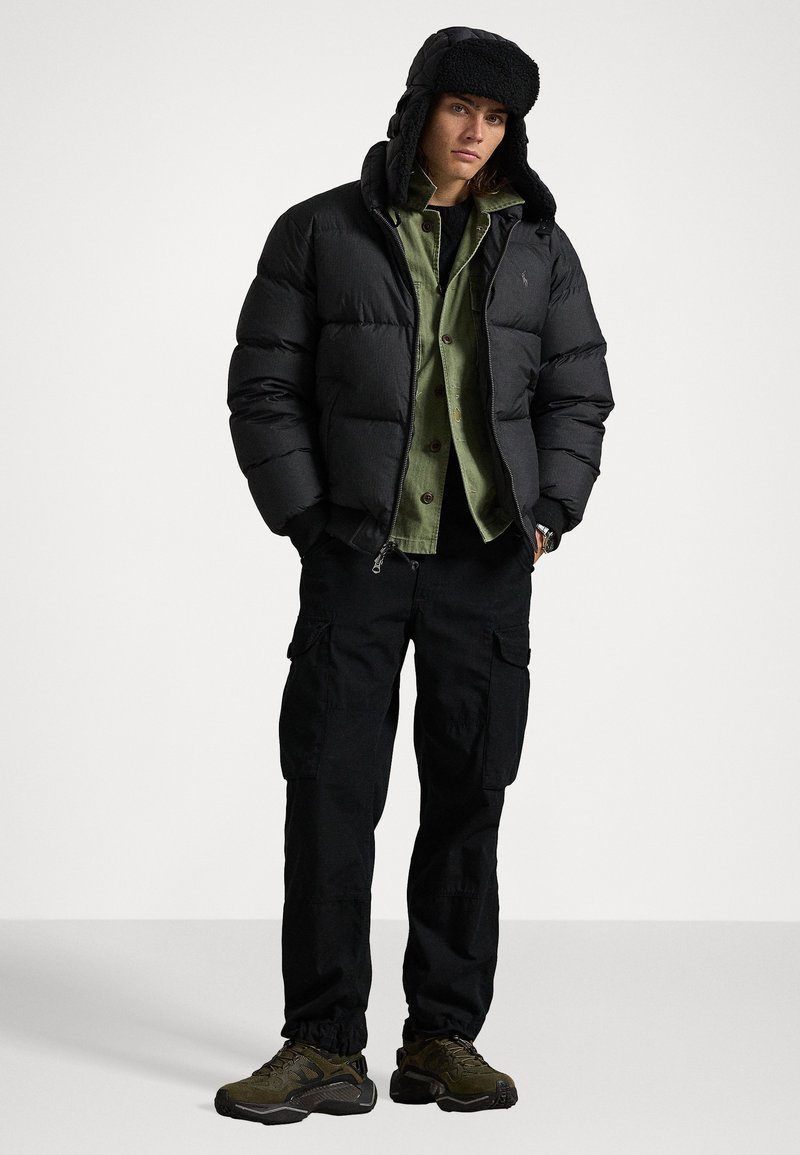 Zwarte pufferjack met een pluizige capuchon, groene button-up shirt eronder, zwarte cargobroek en donkere sportschoenen. Casual buitenstijl.