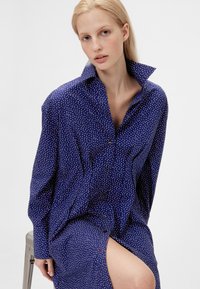 Navy blue shirt-dress with white polka dots, buttoned front, large collar, and side slit. Lightweight fabric with a smooth texture.