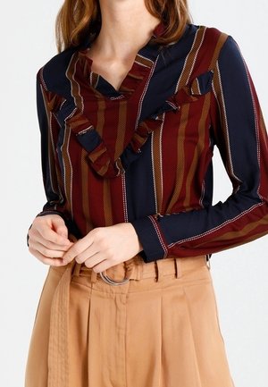 Kobieta nosząca pasiastą, granatową, burgundową i brązową bluzkę z falbankami, wpuszczoną w beżowe spodnie z wysokim stanem, z dopasowanym paskiem i srebrną sprzączką w kształcie pierścienia.