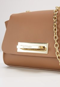 Borsa a tracolla in finta pelle marrone con catena dorata e chiusura rettangolare con la scritta "ZAC POSEN" incisa. Texture liscia, design minimalista.