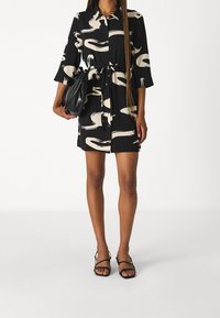 Abito nero con motivo astratto beige, apertura con bottoni, cintura in vita e maniche a tre quarti. Gli accessori includono una borsa nera e sandali con cinturini.