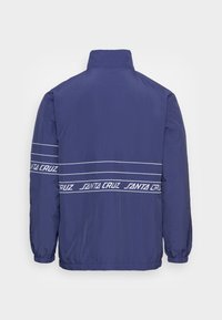Chaqueta cortaviento de color marino con un cuello alto, puños elásticos y un logo en blanco "SANTA CRUZ" en rayas a lo largo de la espalda y las mangas.