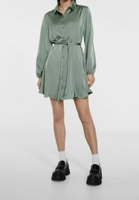 Robe chemise en satin vert avec des manches longues, un col, une fermeture à boutons, une ceinture à la taille et une jupe évasée. Assortie à des chaussures noires épaisses et des chaussettes blanches.