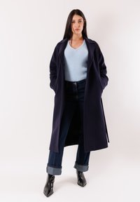 Donna che indossa un lungo cappotto scuro sopra un maglione azzurro chiaro, jeans scuri con risvolto e stivaletti neri con punta, in piedi con le mani nelle tasche.