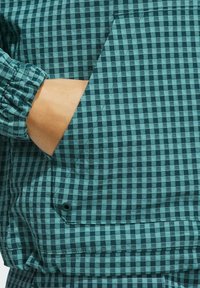 Teal og mørkegrøn ternet stof med lomme detaljer og elastisk ærmekant. Teksturen virker glat og let.