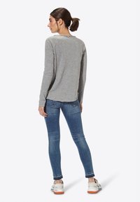 Maglia grigia a manica lunga con scollatura rotonda e orlo grezzo, abbinata a jeans skinny blu. La maglia presenta un discreto dettaglio con bottoni sul retro.