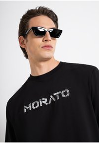 Camiseta de algodón negra con la palabra "MORATO" en una tipografía de patrón único. El modelo lleva gafas de sol negras angulares. Textura suave.