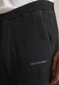 Schwarze Sweatpants mit geripptem Bund, Seitentaschen und dem weißen "MARC O'POLO DENIM" Branding nahe der Tasche. Weiche Textur.