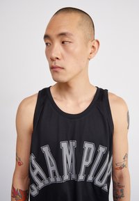 Juodas tinklinis sportinis tank topas su ryškiu "CHAMPION" užrašu pilkomis raidėmis ant krūtinės. Rodymos matomos tatuiruotės ant rankos.