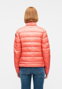 Femme aux cheveux rouges raides mi-longs, portant une doudoune corail vif et un jean bleu, vue de dos sur un fond blanc uni.