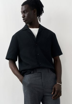 Mann mit Dreadlocks trägt ein schwarzes kurzärmeliges Hemd über einem weißen T-Shirt und graue Hose, steht mit einer Hand in der Tasche und blickt nach links.