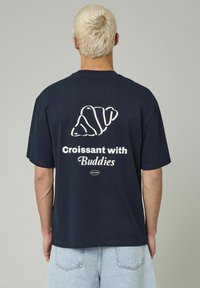 T-shirt en coton bleu marine avec un graphique de croissant blanc et le texte "Croissant avec des amis" au dos. Coupe décontractée, manches courtes.