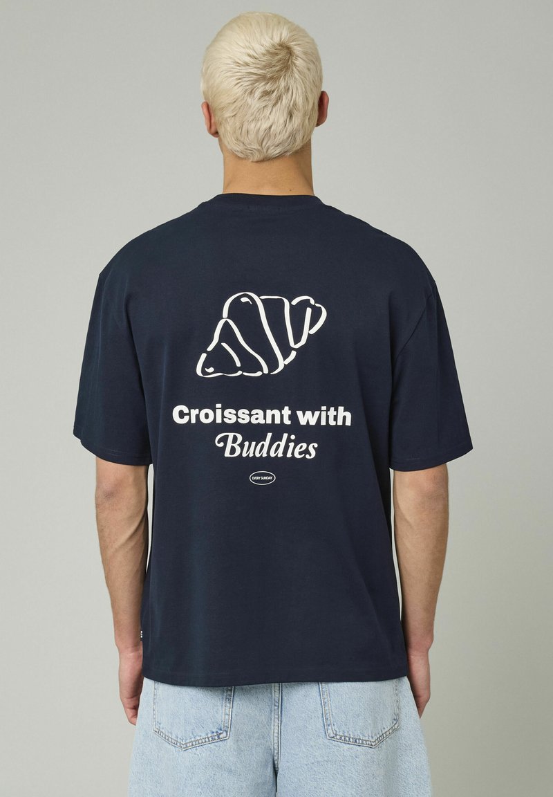 T-shirt en coton bleu marine avec un graphique de croissant blanc et le texte "Croissant avec des amis" au dos. Coupe décontractée, manches courtes.