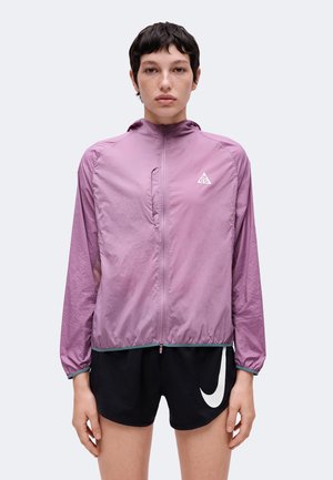 Person med kort mørkt hår iført en lyse lilla lynlåstrøje og sorte shorts med hvidt Nike swoosh-logo.
