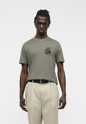 Homme aux cheveux tressés portant un t-shirt graphique vert olive rentré dans un pantalon plissé beige avec ceinture noire, debout devant un fond uni.