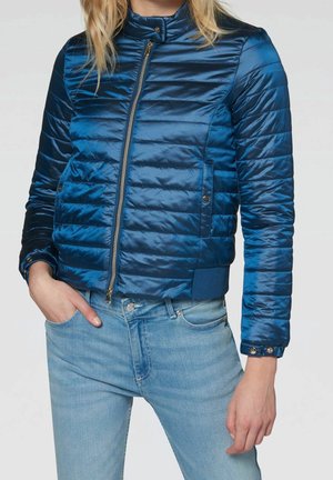 Femme portant une veste matelassée bleue brillante avec une fermeture éclair et un jean bleu clair, main dans la poche, debout devant un fond uni.