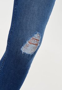 Jeans de mezclilla azul con un desgarrón deshilachado en el muslo, exponiendo la piel debajo de los hilos de tela rasgados.
