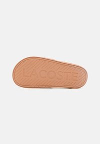 Semelle de chaussures Lacoste, présentant un motif texturé et un logo 'LACOSTE' en relief, d'une couleur brun doux avec des bords arrondis.