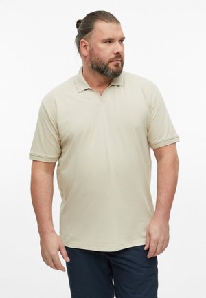 Mężczyzna z brodą i włosami związanymi w koczek nosi jasnobeżową koszulkę polo z krótkim rękawem i ciemne spodnie, stojąc na tle jednolitego białego tła.